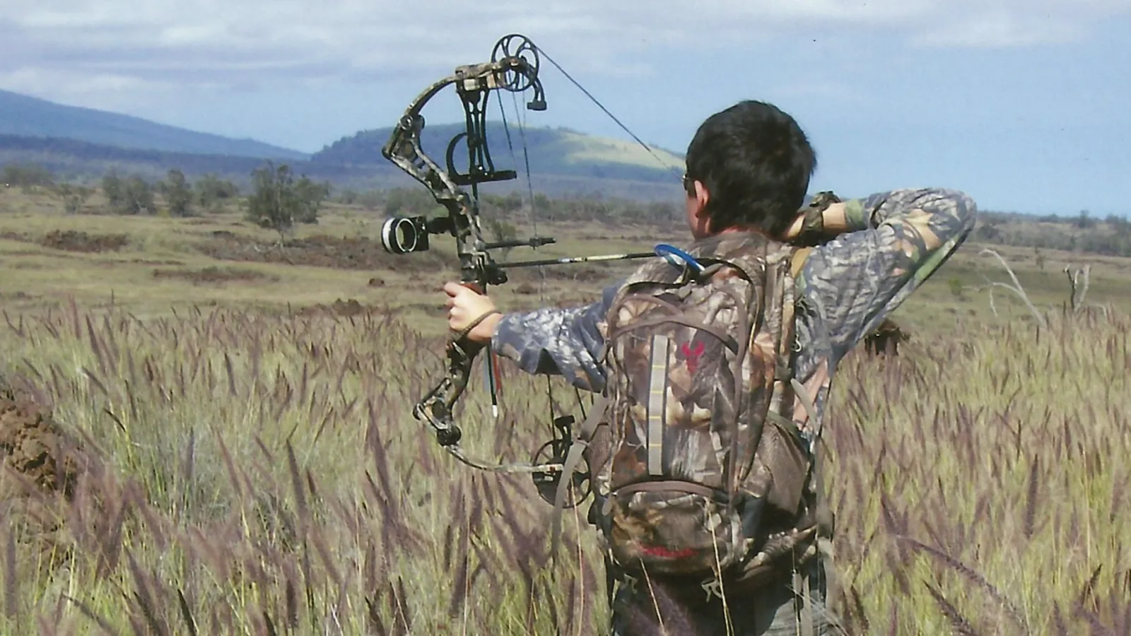 Tyler drawing a bow while hunting in an open field in Hawai'i